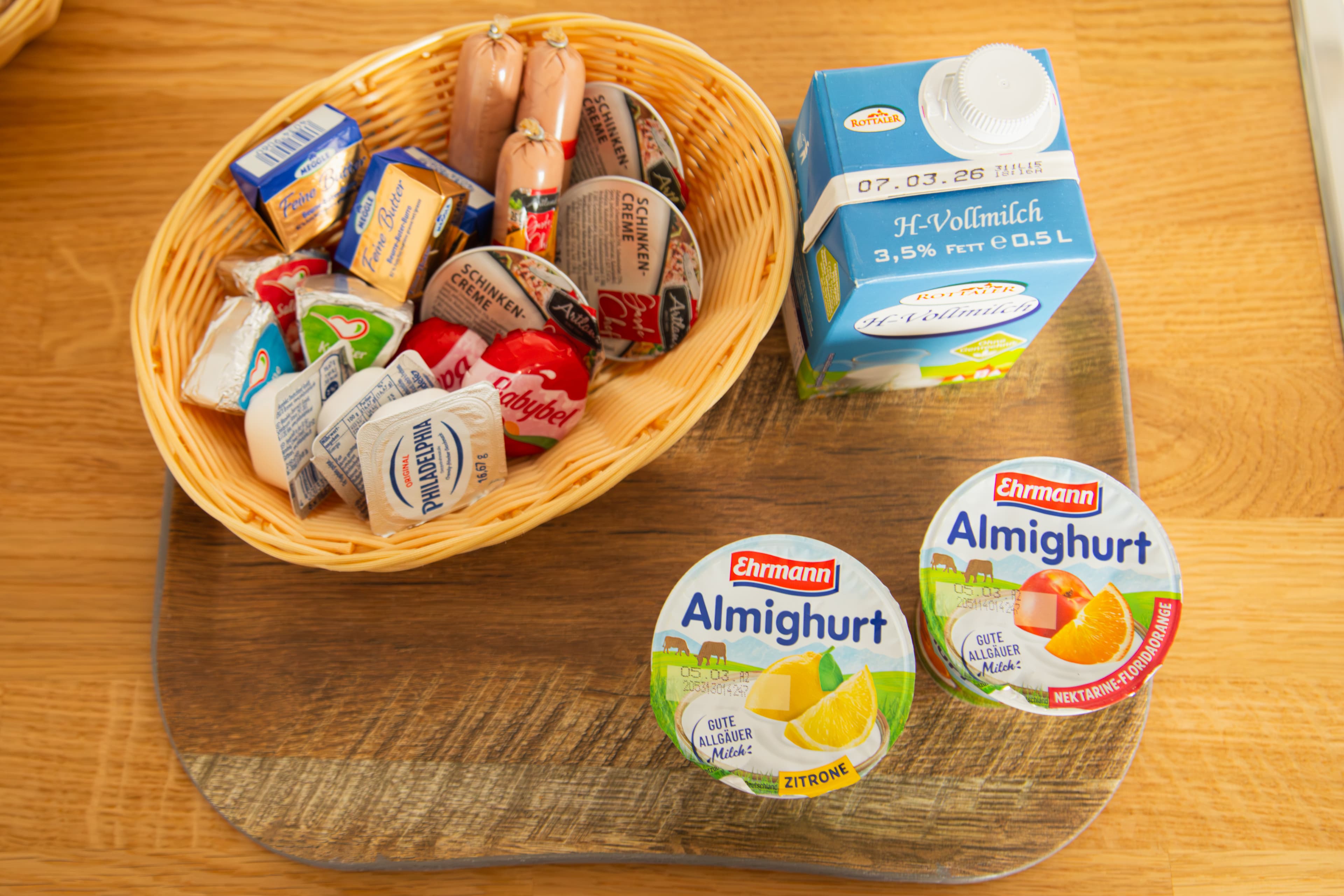 Korb mit Wurst- und Käseaufschnitt, Vollmilch und zwei Almighurt-Bechern auf Holzbrett